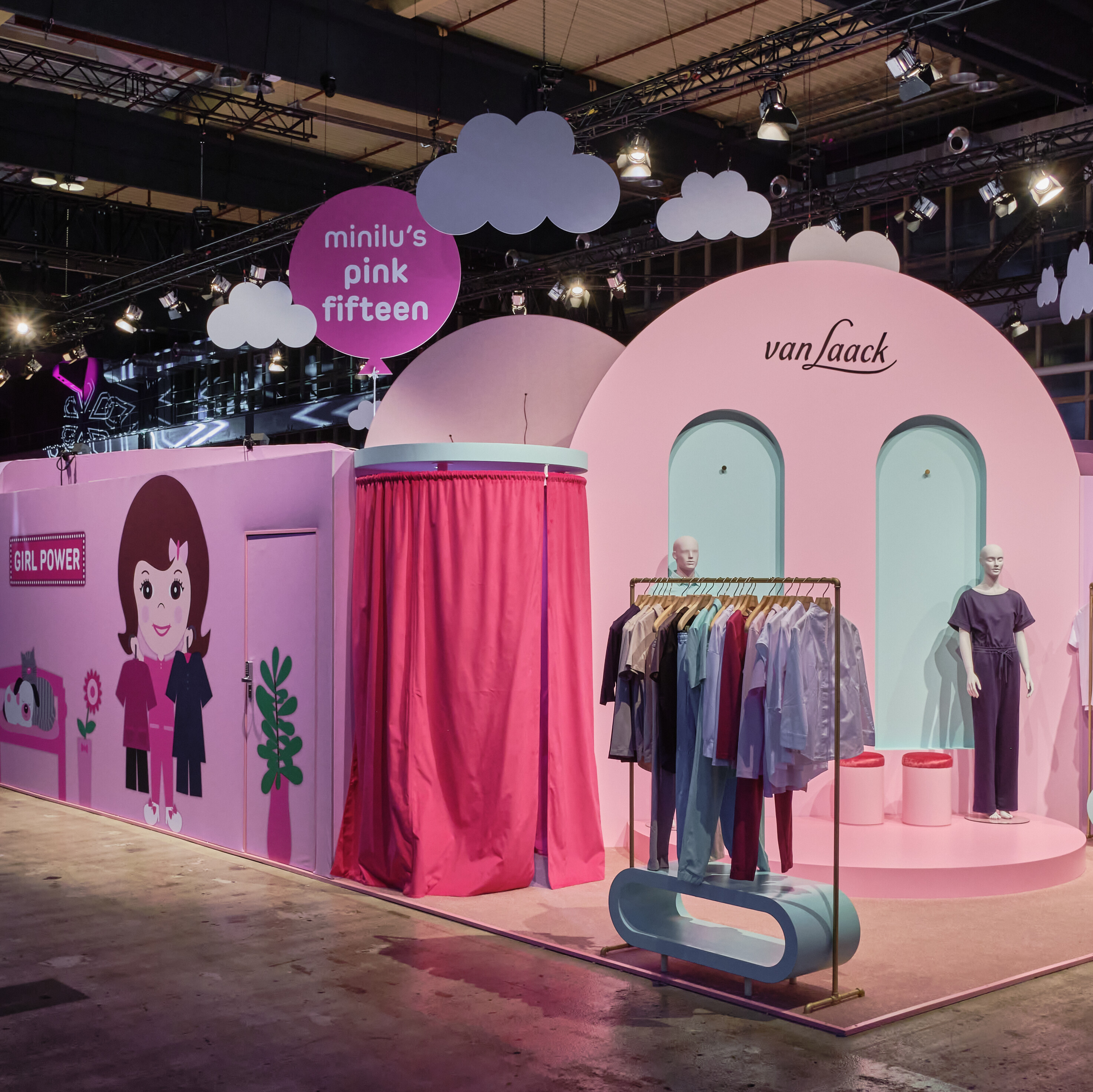 Eine Innenansicht des Events "minilu's Wonderland" mit rosafarbenen, hausförmigen Ständen. Ein Stand zeigt den Schriftzug "van Jaack" und Kleiderstangen mit Kleidung, während ein anderer Stand mit dem Slogan "Girl Power" und einer Comic-Figur versehen ist. Über den Ständen schweben stilisierte Wolken aus Pappe. Die Szene ist hell ausgeleuchtet. Eine Innenansicht des Events "minilu's Wonderland" mit rosafarbenen, hausförmigen Ständen. Ein Stand zeigt den Schriftzug "van Jaack" und Kleiderstangen mit Kleidung, während ein anderer Stand mit dem Slogan "Girl Power" und einer Comic-Figur versehen ist. Über den Ständen schweben stilisierte Wolken aus Pappe. Die Szene ist hell ausgeleuchtet.