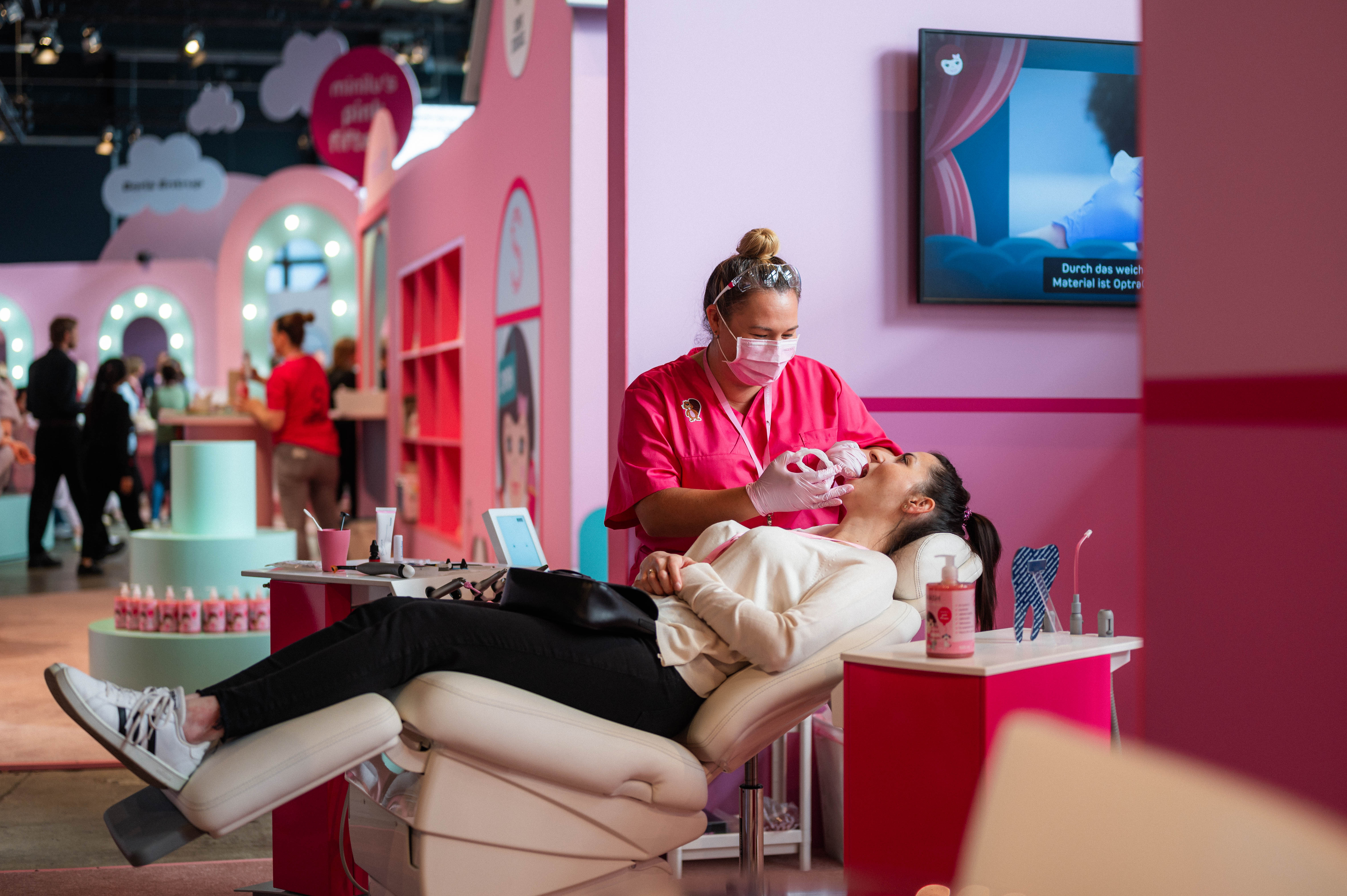 Eine Frau mit Maske und pinker Arbeitskleidung beugt sich über eine Person, die in einem Behandlungsstuhl liegt. Im Hintergrund ist ein großer Bildschirm zu sehen, der eine Produktpräsentation zeigt. Die Szene ist Teil des Events, das in rosafarbenen und weißen Tönen gehalten ist. Eine Frau mit Maske und pinker Arbeitskleidung beugt sich über eine Person, die in einem Behandlungsstuhl liegt. Im Hintergrund ist ein großer Bildschirm zu sehen, der eine Produktpräsentation zeigt. Die Szene ist Teil des Events, das in rosafarbenen und weißen Tönen gehalten ist.